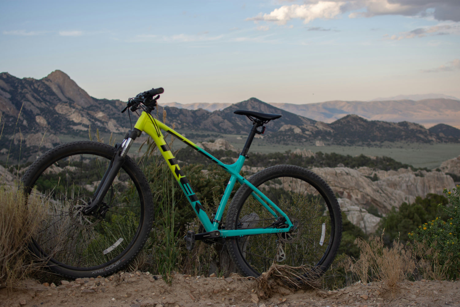 A bright yellow and turquoise hardtail mountain bike stands on a rocky trail with wide mountain views in the background, illustrating the type of used MTBs available on premium UK marketplaces like MyNextBike.co.uk. 
