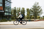 Woman riding a black electric bike with a helmet and backpack on a city street near modern buildings and trees. 