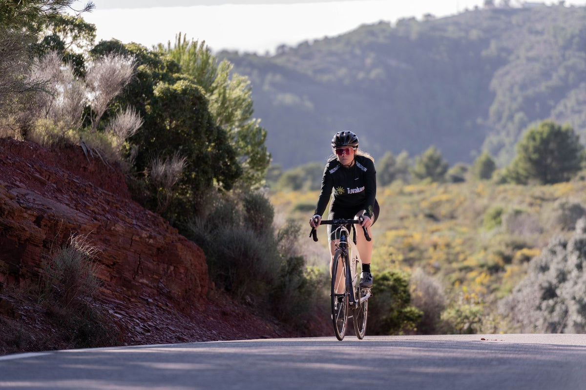 Woman in a helmet riding a drop-bar road bike up a quiet rural road between green hills