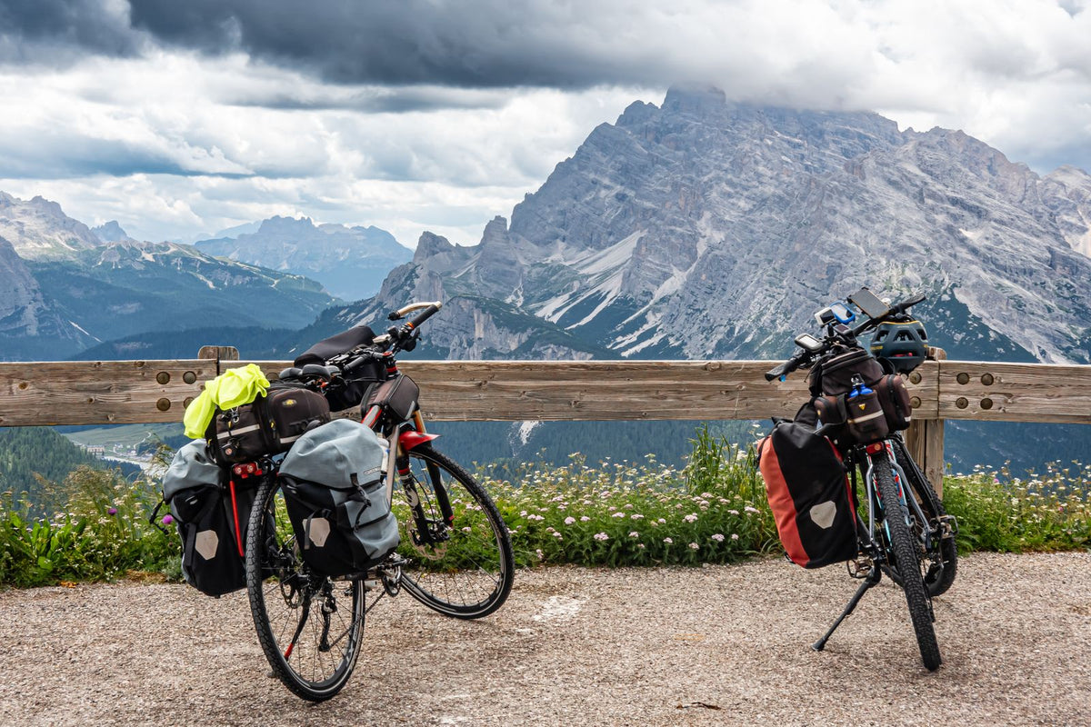 Two loaded touring bikes with panniers and rack bags resting by a wooden barrier in front of a mountain range