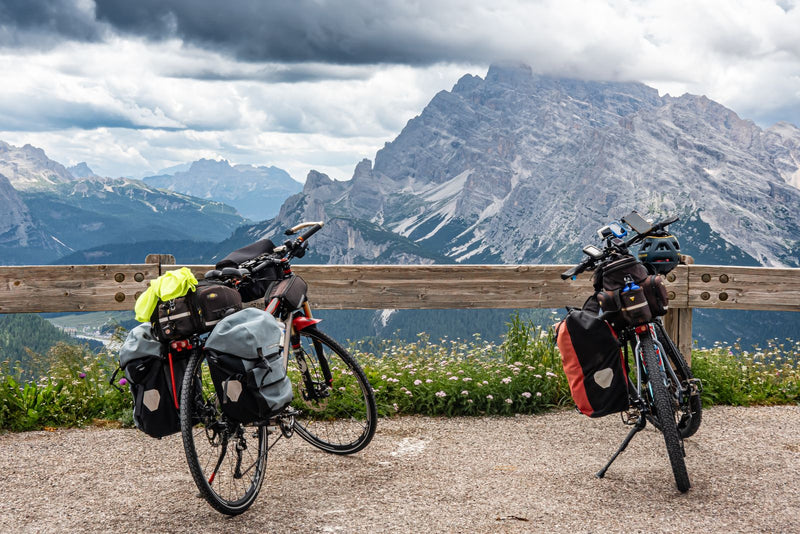 Two loaded touring bikes with panniers and rack bags resting by a wooden barrier in front of a mountain range