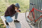 A photographer crouches to capture a shot of a red bicycle on a sunny day.