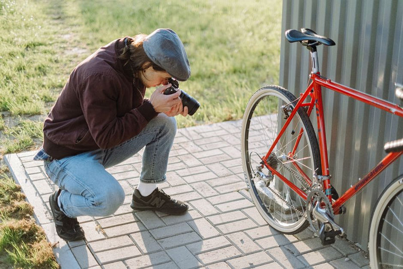 A photographer crouches to capture a shot of a red bicycle on a sunny day.