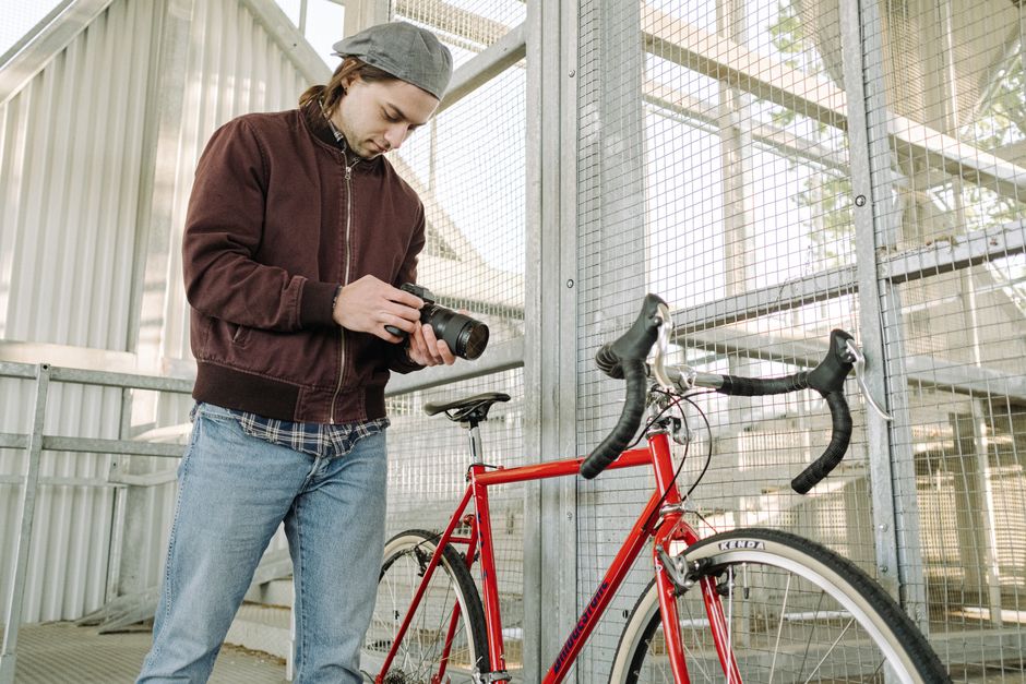 A young man standing beside a red bicycle, inspecting his camera outdoors.