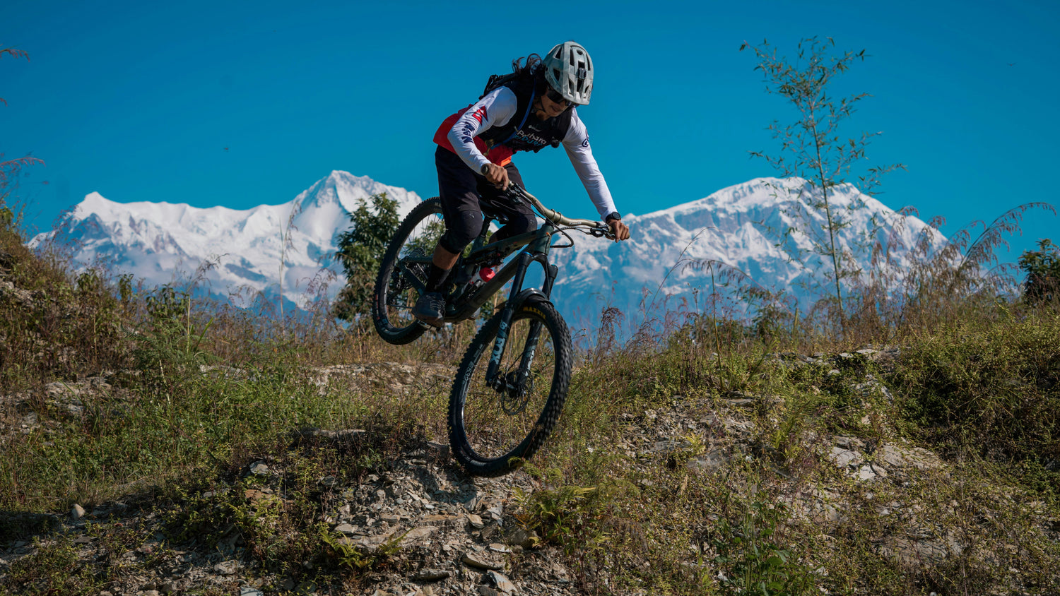Woman on a full-suspension mountain bike jumping over a rocky trail with snow-capped mountains in the background, representing women’s MTB riding and outdoor adventure -- find yours at MyNextBike.co.uk. 