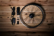 Electric bike conversion kit with hub motor wheel, battery, charger and components laid out on wooden table.